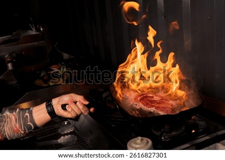 Chef flambéing steak in hot pan with dramatic fire in kitchen setting.