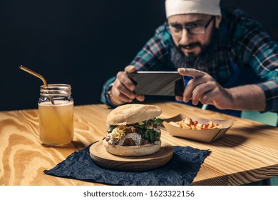 Chef Taking A Photo With The Phone Of His Vegan Burger With Lettuce And Sauce, Fries With Ketchup And Healthy Drink On A Wooden Table, Technology And Gourmet Lifestyle Concept, Selective Focus