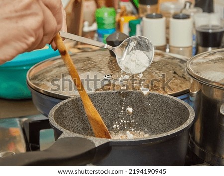 Chef throws flour into pot of bechamel sauce being prepared on gas stove.