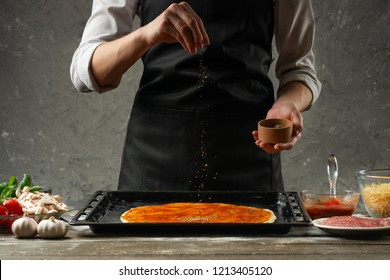 Chef In Uniform Adding Spice To Pizza After Cooking Pizza On The Background Of A Concrete Wall