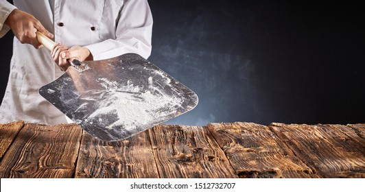 Chef In Uniform Holding An Empty Floured Metal Paddle For Cooking Pizza In A Wood Fired Oven Over A Rustic Table In A Panorama Banner With Copy Space For Food Placement