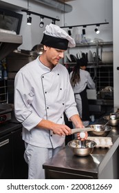 Chef In Uniform Holding Knife Near Raw Egg Above Bowl While Cooking In Kitchen