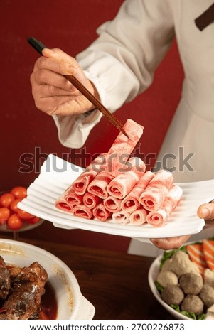 Chef using chopsticks to pick up thinly sliced raw meat rolls from white plate, with hot pot and meatballs visible in background for Asian cooking preparation