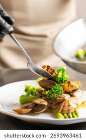 A chef using a spoon to plate a dish of fresh steamed clams with green vegetables like broccoli and asparagus. A close-up shot capturing the final stage of cooking