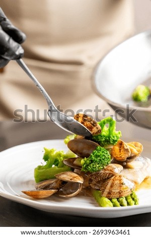 A chef using a spoon to plate a dish of fresh steamed clams with green vegetables like broccoli and asparagus. A close-up shot capturing the final stage of cooking