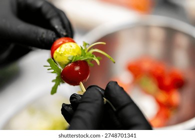 A chef wearing black gloves assembles a Caprese skewer with cherry tomatoes, mozzarella, and fresh arugula over a metal bowl.