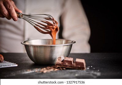 Chef whisking melted chocolate in a stainless steel mixing bowl using an old vintage wire whisk in a close up on his hand - Shutterstock ID 1327917536