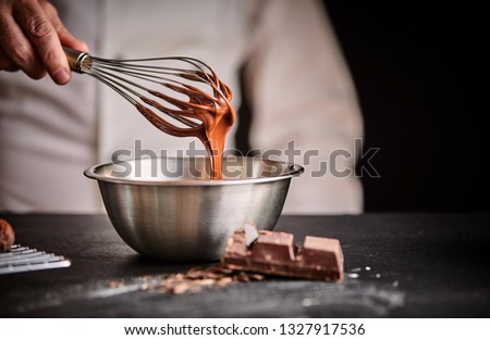 Chef whisking melted chocolate in a stainless steel mixing bowl using an old vintage wire whisk in a close up on his hand