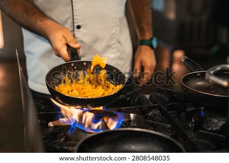 A chef in a white jacket flips creamy orange pasta in a black skillet over a gas flame in a professional kitchen, with warm low lighting and shallow depth of field.