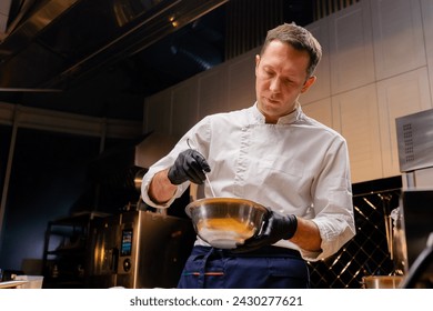 a chef in a white jacket in the kitchen stirs the contents in a pan with a whisk while holding it in his hands - Powered by Shutterstock