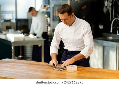 a chef in a white jacket in the kitchen at the table sharpens a knife against the background his assistant