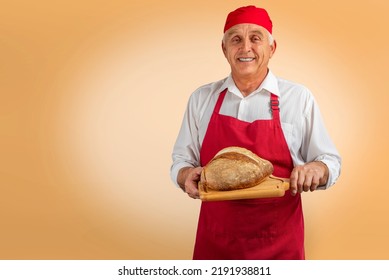 Chef-cooker In Chef's Hat And Apron Working In Bakery, Holding French Bread Board With Bread. Senior Professional Baker Man Wearing Chef's Outfit. Character Kitchener, Pastry Chef For Advertising.