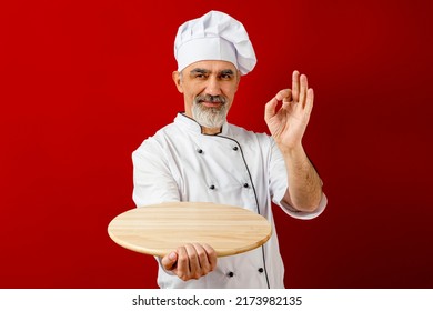 Chef-cooker In A Chef's Hat And Jacket Gesturing Okay, Holding An Empty Wooden Tray For Mock Up. Senior Professional Baker Man Wearing A Chef's Outfit. Character Kitchener, Pastry Chef For Advertising