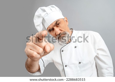 Chef-cooker in a chef's hat and jacket pointing index finger at the camera. Senior suspecting baker man wearing a chef's outfit. Character kitchener, pastry chef for advertising.