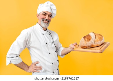 Chef-cooker In Chef's Hat And Jacket Working In Bakery, Holding French Bread Board With Bread. Senior Professional Baker Man Wearing Chef's Outfit. Character Kitchener, Pastry Chef For Advertising.