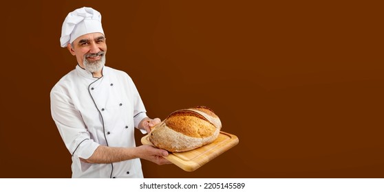 Chef-cooker In Chef's Hat And Jacket Working In Bakery, Holding French Bread Board With Bread. Senior Professional Baker Man Wearing Chef's Outfit. Character Kitchener, Pastry Chef For Advertising.