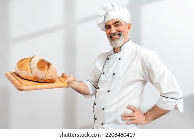 Chef-cooker In Chef's Hat And Jacket Working In Bakery, Holding French Bread Board With Bread. Senior Professional Baker Man Wearing Chef's Outfit. Character Kitchener, Pastry Chef For Advertising.