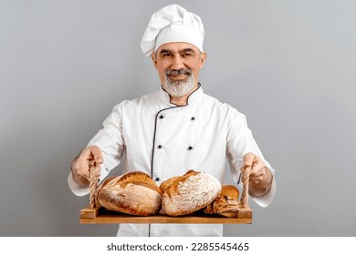 Chef-cooker in a chef's hat and jacket working in bakery, holding tray with bread and rolls. Senior professional baker man wearing a chef's outfit. Character kitchener, pastry chef for advertising. - Powered by Shutterstock