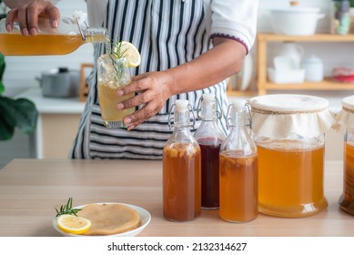 Chef's hand holds a bottle and a glass of Homemade fermented kombucha tea, variety of flavors in bottles and glass jars arranged mix with fruit, scoby on wooden table. Healthy natural probiotic drink.