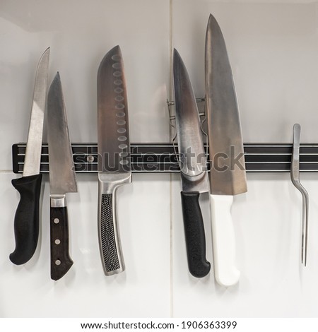Chef's knives and tweezer on a magnetic holder. A set of knives for cutting food. Kitchen utensils. Magnetic kitchen knife holder closeup. Multiple professional knives and tweezer.