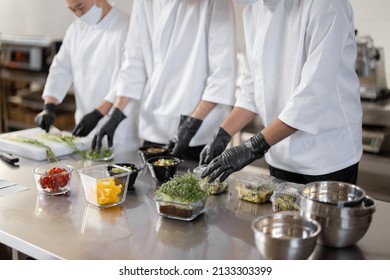 Chefs prepare takeaway food, mixing ingredients in disposable dish at professional kitchen. Concept of a dark kitchen for cooking for delivery during pandemic. Cropped view without face