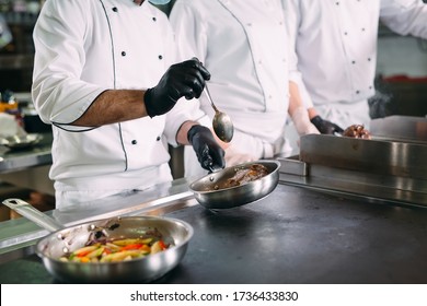 Chefs In Protective Masks And Gloves Prepare Food In The Kitchen Of A Restaurant Or Hotel