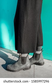 Chelsea Boots Classic Black Leather Rubber Sole. Focus On Legs Of Hipster Woman Wearing Large Oversized Wide Leg Black Trousers. Shot On Street With Shadows On Pavement.