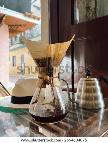 A Chemex coffee maker with a filter is brewing coffee on a reflective counter. In the background, a Panama hat and a gooseneck kettle complete the cozy, artisanal cafe scene