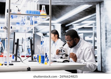 Chemical engineer operating compound microscope, doing detailed sample observation in controlled laboratory environment. African american man studies tissue under optical device in biotechnology lab