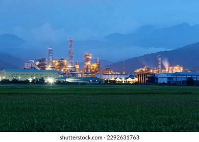 A chemical factory in green rice paddies before distant mountains with lights glowing and chimneys smoking in blue twilight. Factory pipes polluting air at silent dusk, a serious environmental issue - Shutterstock ID 2292631763