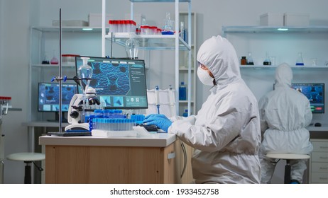 Chemist in ppe suit typing on computer checking virus development in equipped laboratory. Team doctors working with various bacteria, tissue and blood samples, pharmaceutical research for antibiotics