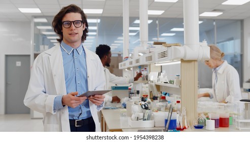 Chemist using tablet computer in lab talking on camera. Portrait of young male scientist in white coat giving interview standing in modern research laboratory - Powered by Shutterstock