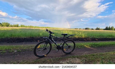 Cheremshan, Tatarstan, Russia, June 07, 2021: A Bicycle Stands On A Dirt Road Against The Background Of Pouring Rain And A Rainbow. Outskirts Of The Village.