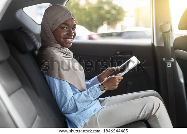 Cherful Black Woman In Hijab Using Digital\
Tablet In Car, Sitting On Backseat And Smiling At Camera, Positive\
Islamic Businersswoman Enjoying Modern Technologies And Comfortable\
Transportation