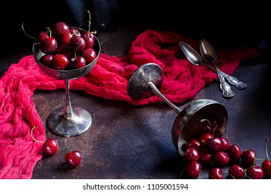 Cherries In A Silver Vase. Red Berries In Droplets Of Water. Antique Silverware. Red Runner. Cherries Scattered On The Table. Dark Photo. Selective Focus.