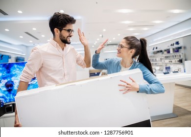 Cherry Young Couple Giving High-five While Carrying TV. Tech Store Inteior.