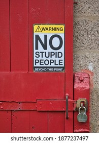 Cheshire, UK - Dec 9 2020: A humorous safety sign saying "Warning! No stupid people beyond this point" fixed to a red, wooden door locked with a padlock and hasp.