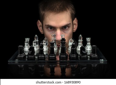 Chess Master staring at you with intense eyes behind glass chess pieces on a glass chessboard with a reflection isolated on a black background