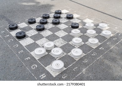 A chessboard on asphalt with white and black pieces for playing checkers and competing in a battle on the square in front of the City Administration building near the park. Street chess game - Shutterstock ID 2179380401