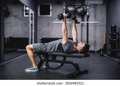 Chest and arm workout at sports bench with dumbbells. A man in sportswear lies on a sports bench and lifts dumbbells, training with a load. Strength, powerful body and physical endurance