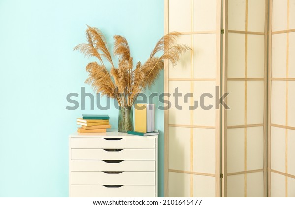 Chest of drawers with vase, books and folding screen\
near blue wall