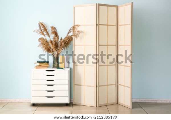 Chest of drawers with vase, books and folding screen\
near blue wall