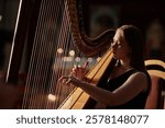 Chest up shot of concentrated young woman playing harp delicately plucking strings of instrument while performing classical music in cinematic side view light on stage