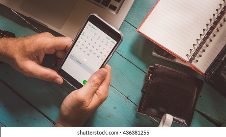 Chiang Mai, Thailand - June 13, 2016 A Man Holding A Samsung Galaxy J5 Phone, Open Is The Calendar App. Also On The Table Are His Wallet And Laptop