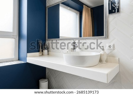 A chic bathroom featuring a modern, round white sink, a clean white countertop, a framed mirror, and stylish white herringbone tiles against a deep blue wall