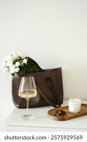 A chic composition featuring a stylish designer bag with fresh flowers , a beautiful glass of wine, chocolote, candle on wooden tray. High quality photo