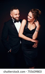 Chic, Elegant Couple: Handsome Man With Beard In Tuxedo With Amazing Woman With Blonde Updo Hair, Wearing Silky Black Dress And Luxurious Jewelry, Posing In Dark Studio