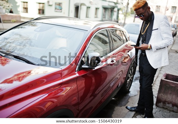 Chic handsome african american\
man in white suit against red luxury car with mobile phone at\
hand.