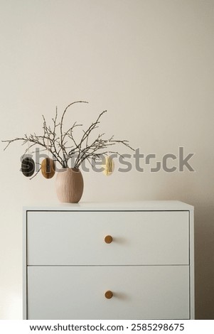 Chic minimalist easter decor features dry branches with craft eggs elegantly arranged in a simple vase, easter rabbit set a top a modern dresser. High quality photo