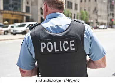 A Chicago city policeman standing on the road on a sunny day wearing a bullet proof vest mentioning Police on his body. On the background, cars and tall buildings are seen.